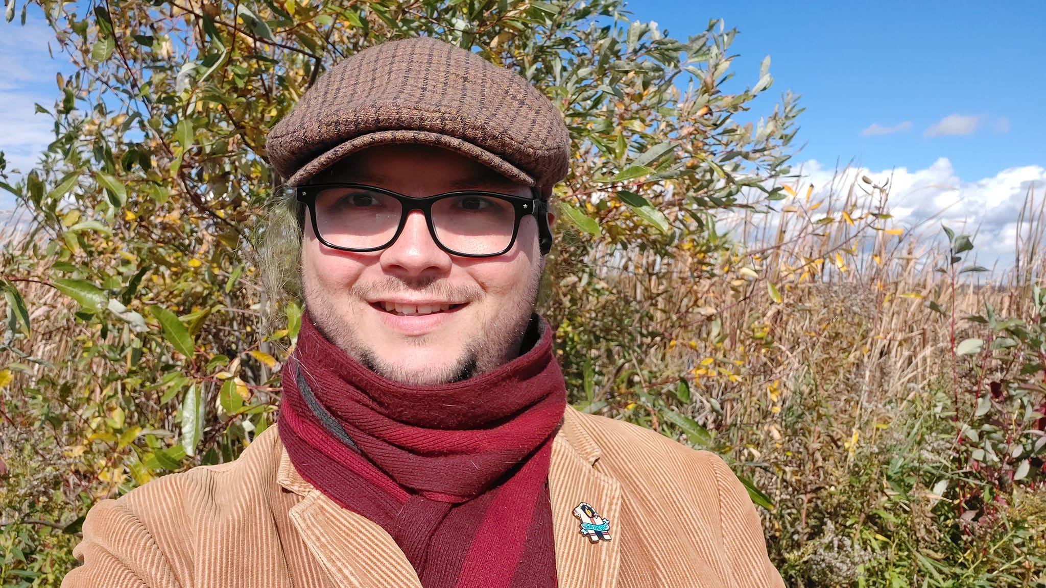 A man with long green hair wearing a brown corderoy jacket and red scarf satands in front of a wetland